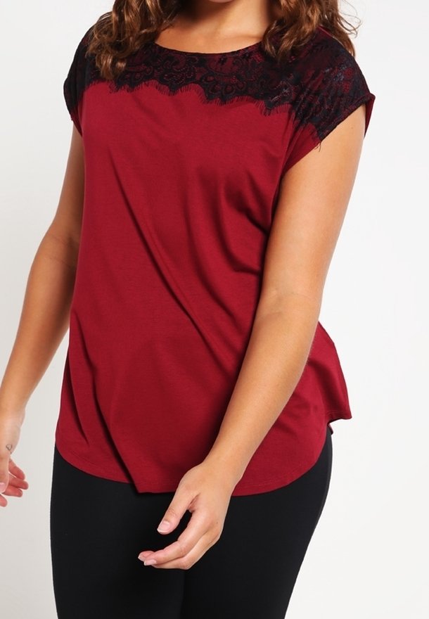 Kvinna klädd i en kortärmad maroon topp med svart spetsdetalj över axlarna, ihop med svarta byxor mot en vit bakgrund.