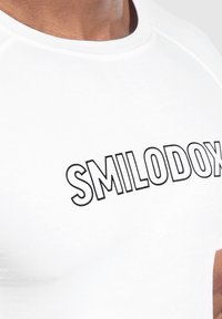 Valkoinen istuva paita, joka on valmistettu sileästä kankaasta. Paidassa on musta "SMILODOX" -teksti rinnan kohdalla, pyöreä kaula-aukko ja lyhyet hihat.