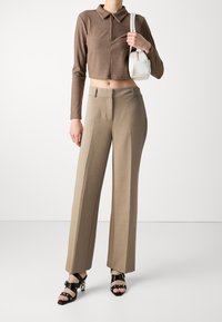 Hnědý žebrovaný polo top s dlouhým rukávem, spárovaný s béžovými širokými kalhotami s vysokým pasem a černými sandály na tenké šňůrky, držící bílou kabelku.