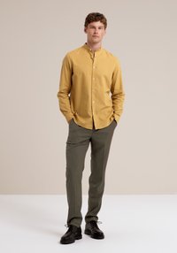Cămașă din in de culoare muștar cu guler mandarin, mâneci lungi și nasturi în față, asortată cu pantaloni verzi măslinii și pantofi negri.
