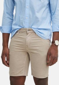 Shorts de algodón beige con un corte ajustado, que cuentan con cierre de botón y cinco bolsillos. Combinados con una camisa de manga larga azul clara.