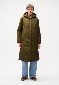 Olijfgroene gewatteerde jas met een capuchon, koordjes, twee zijzakken en een lange, rechte snit. Draagt over losse blauwe jeans en bruine sneakers.