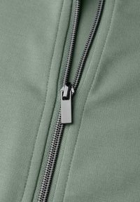 Tessuto verde con cerniera a zip in metallo dotata di cursore nero lucido, che mette in risalto la superficie testurizzata e i dettagli delle cuciture.