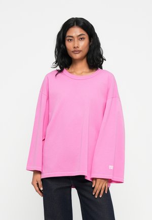 Übergrößer, rosa Sweatshirt aus Baumwolle mit weitem Ausschnitt und langen Ärmeln. Mit weißem Logo-Detail am Saum. Getragen mit dunklen Jeans.