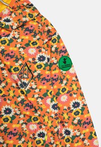 Oranje jas met bloemenpatroon, voorzien van zwart diamantlogo en groene cirkelpatch. Gemaakt van een gladde stof met levendige kleuren.