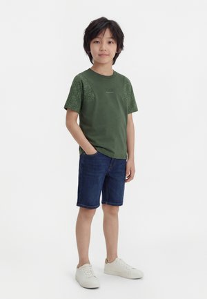 Garçon aux cheveux noirs portant un T-shirt vert à motifs, un short en jean bleu et des baskets blanches, debout avec une main dans la poche.