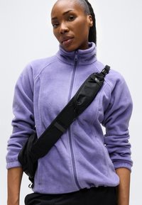 Γυναίκα που φοράει ένα φωτεινό μωβ φλις μπουφάν και μια μαύρη τσάντα χιαστί μπροστά σε απλό ανοιχτό φόντο.