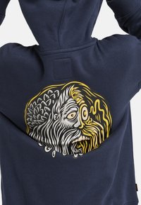 Un hoodie bleu marine avec un grand graphique au dos représentant un visage stylisé en blanc, noir et jaune, avec des cheveux fluides et des détails.