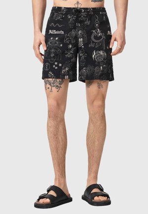 Mand iført sorte shorts med hvide abstrakte og blomsterprint samt sorte sandaler med dobbelt rem, stående mod en ensfarvet baggrund.