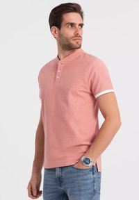 Růžová bavlněná polo košile s henley límcem, tříknoflíkovou légou a bílými akcenty na rukávech, doplněná modrými džínami.