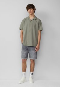 Kortekorte, lichtgroene gestructureerde blouse met knopen aan de voorkant, gecombineerd met opgerolde grijze denim shorts en witte sneakers met zwartgestreepte sokken.