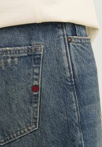 Denim jeans in light blue with visible stitching, a pocket on the back, and a small red detail near the top right of the pocket.