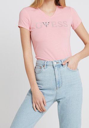 Roze T-shirt met korte mouwen en V-hals, voorzien van "GUESS" gedrukt in zilveren rhinestones; gecombineerd met hoge lichtblauwe jeans.