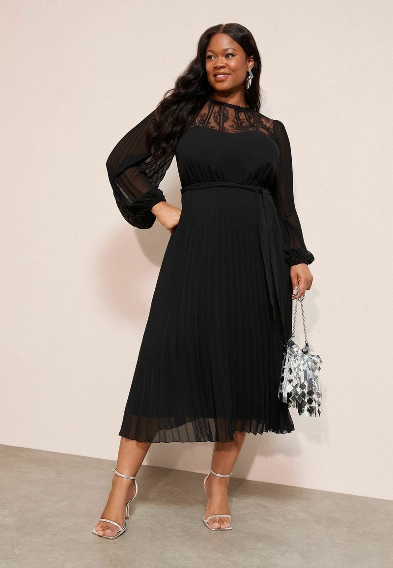 Fekete plisszírozott ruha áttetsző hosszú ujjakkal és csipkebodóval. Ezt egy flitteres ezüst táska és ezüst pántos magassarkú cipő kíséri.