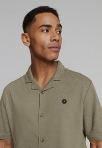 Korte mouwen olijfgroene polo met een gestructureerde stof, twee knopen en een zeshoekig zwart embleem op de linkerborst.