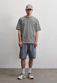 Ung mann iført grå oversized t-skjorte, matchende grå shorts, lysegrå caps, hvite sokker og beige sneakers som står mot enkel bakgrunn.