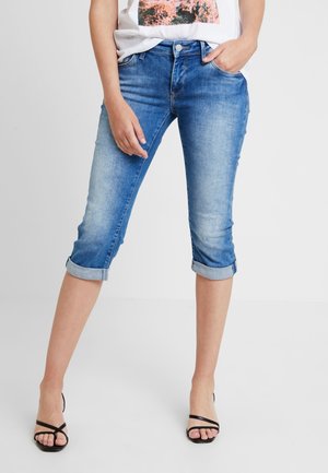Mujer con pantalones capri vaqueros azules remangados, camiseta blanca con diseño gráfico y sandalias negras con tacón y tiras, de pie con una mano en el bolsillo.