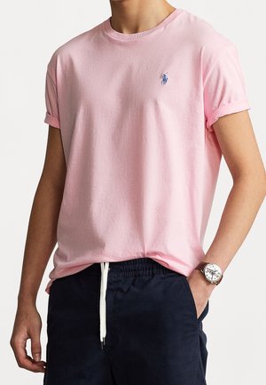 Homme portant un t-shirt rose clair à manches retroussées et un short bleu foncé avec cordon de serrage, avec une montre au poignet gauche.