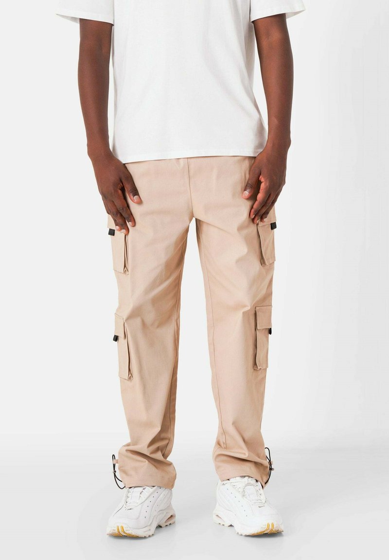 Beige cargobroeken met meerdere zijzakken, trekkoordmanchetten en een gladde textuur, gecombineerd met een wit T-shirt met ronde hals.