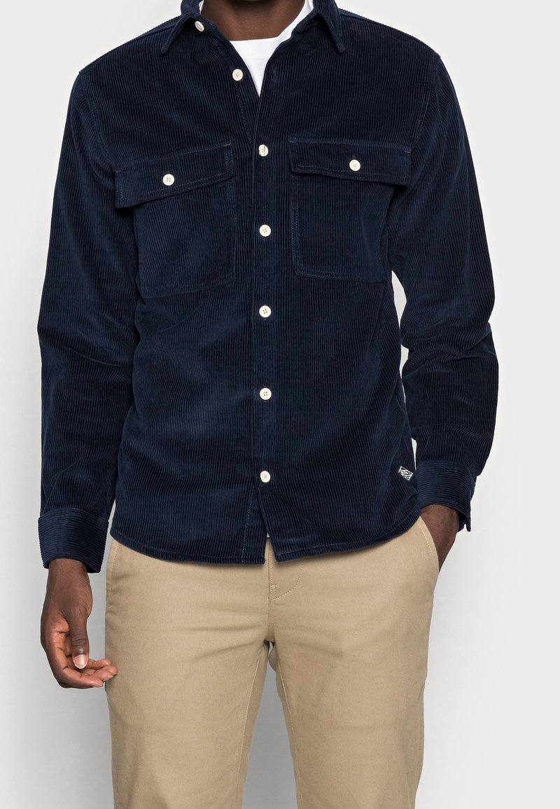 Marinblå kordflanellskjorta med två framfickor, knäppning med knappar och vita knappar. Har långa ärmar med manschetter och rundad nederkant.