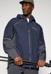 Navy and gray waterproof jacket with a hood, zip closure, and logo detail. Lightweight fabric with contrasting panel accents.
