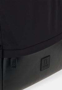 Svart väska med en slät nylonöverdel och en texturerad läderbotten. Har en logotypfärg och en liten märkestämpel. Minimalistisk design.