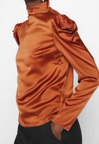 Hochkragenoberteil aus rostfarbenem Satin mit langen Ärmeln, gepufften Schultern und glatter Textur. Besitzt eine figurbetonte Silhouette und dezente Nahtdetails an den Seiten.
