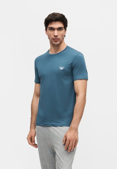 Άνδρας που φοράει μπλε κοντομάνικο μπλουζάκι με μικρό λογότυπο στο στήθος και ανοιχτό γκρι παντελόνια, στέκεται μπροστά σε απλό φόντο.