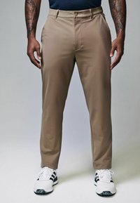 Khakifarbene Chinos mit maßgeschneidertem Schnitt, flachem Bund, Gürtelschlaufen und Seitentaschen. Getragen mit weißen und schwarzen Sportschuhen.