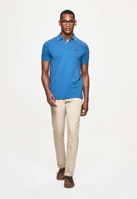 Modrá polokošile s kontrastním límcem, béžové chinos kalhoty a hnědé boty. Model nosí brýle a stojí proti neutrálnímu pozadí.