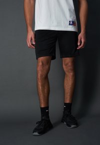 Pantaloni sportivi neri e una t-shirt bianca. La maglietta presenta un piccolo logo; i pantaloni hanno una trama liscia e un design semplice. Le sneakers nere completano il look.