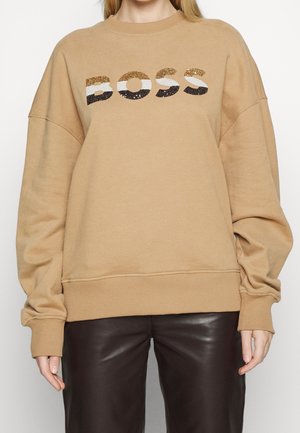 Personne portant un sweat beige avec "BOSS" en sequins dorés, blancs et noirs, associé à un pantalon en cuir marron foncé.