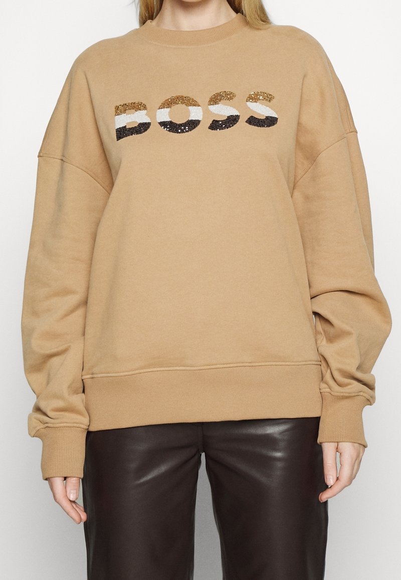 Personne portant un sweat beige avec "BOSS" en sequins dorés, blancs et noirs, associé à un pantalon en cuir marron foncé.
