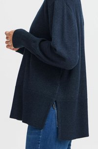 Dunkelblauer Strickpullover mit langen Ärmeln, gerippten Bündchen und Saum, seitlichen Schlitzen, weicher Textur und lockerer Passform, kombiniert mit blauen Jeans.