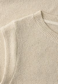 Nærbillede af beige strikket sweaterstof med ribstrikket rund halsudskæring og tekstureret strikkemønster.