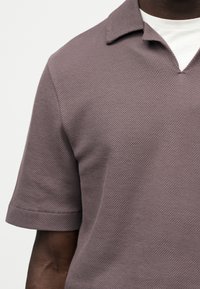 Κοντινό πλάνο ενός ατόμου με σκούρο δέρμα που φοράει υφασμένο κοντομάνικο πόλο πουκάμισο σε αποχρώσεις του μωβ πάνω από λευκό τοπ με στρογγυλή λαιμόκοψη.