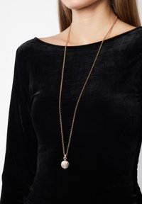 Collar de cadena dorada larga con un colgante redondo que presenta una forma ovalada texturizada y un patrón en zigzag, usado sobre una blusa de terciopelo negra.