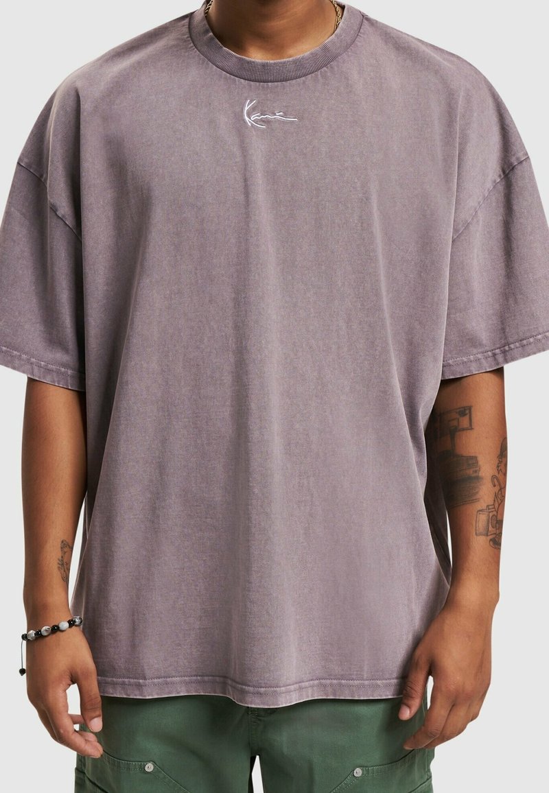Personne portant un T-shirt violet délavé ample avec un petit logo blanc en écriture sur la poitrine, un pantalon vert, des tatouages visibles sur les bras et un bracelet de perles noir et blanc.