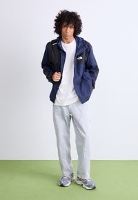 Jūras spēku vēja jaka ar melniem akcentiem, kopā ar vienkāršu baltu T-kreklu, gaiši ziliem džinsiem un rakstainām sporta apavām.