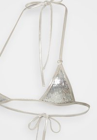 Bikini gornji dio u obliku trokuta prekriven srebrnim šljokicama s tankim, sjajnim trakama. Ima podesive vezice za prilagodbu i reflektirajući teksturu.