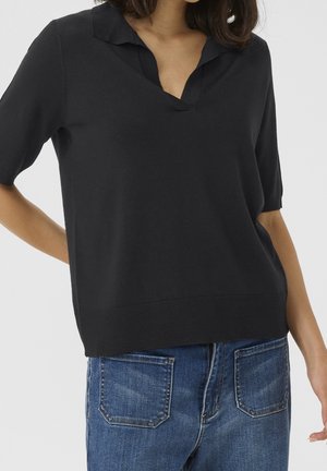 Kvinde iført en sort strikket top med korte ærmer, krave og V-udskæring, kombineret med blå denimjeans med forlommer.