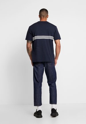 Homme portant un t-shirt bleu marine avec trois rayures blanches horizontales, un jean bleu foncé, des chaussettes blanches et des baskets noires, debout face à l'arrière.