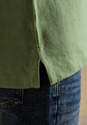 Grüner Baumwollshirt-Saum mit Seitenschlitz über blauen Jeans, zeigt Stoffstruktur und Nahtdetails.