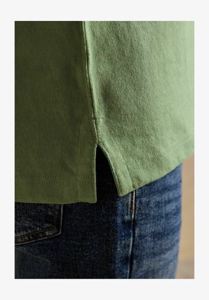 Grüner Baumwollshirt-Saum mit Seitenschlitz über blauen Jeans, zeigt Stoffstruktur und Nahtdetails.