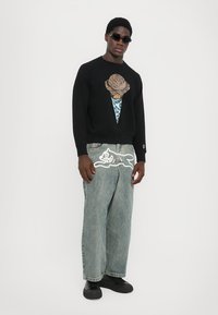Man in een zwart sweatshirt met een ijscomonsterafbeelding, ruime lichtblauwe jeans met een wit cartoonontwerp, zwarte schoenen en zonnebril, staand.