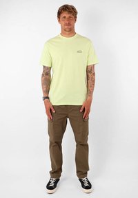 T-shirt en coton vert clair à manches courtes avec un petit logo imprimé sur la poitrine, associé à un pantalon cargo marron et des baskets noires.