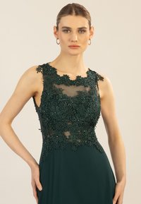 Smaragdgrünes Kleid mit floralem Spitzen-Oberteil und transparenter Ausschnitt. Das glatte Stoffoberteil geht in einen fließenden Rock über; filigrane Perlenverzierungen sind sichtbar.