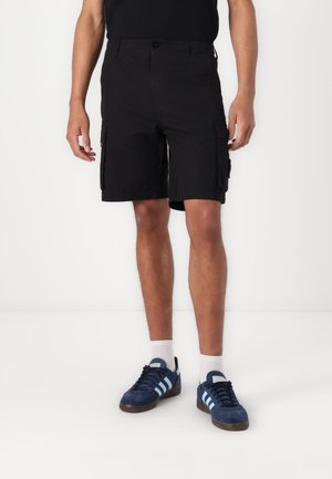 Shorts cargo noirs avec poches latérales, fabriqués à partir d'un tissu léger. Associés à des baskets navy et des chaussettes blanches. Design épuré et décontracté.