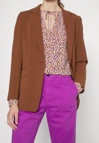 Blazer marron sur un chemisier à motifs multicolores avec un col lacé, assorti de pantalons roses vifs. Comprend des manches longues et une coupe ajustée.