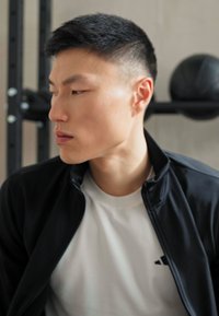 Black zip-up jacket over a white T-shirt, featuring a small black adidas logo. Close-up profile view, with short dark hair and clean skin.